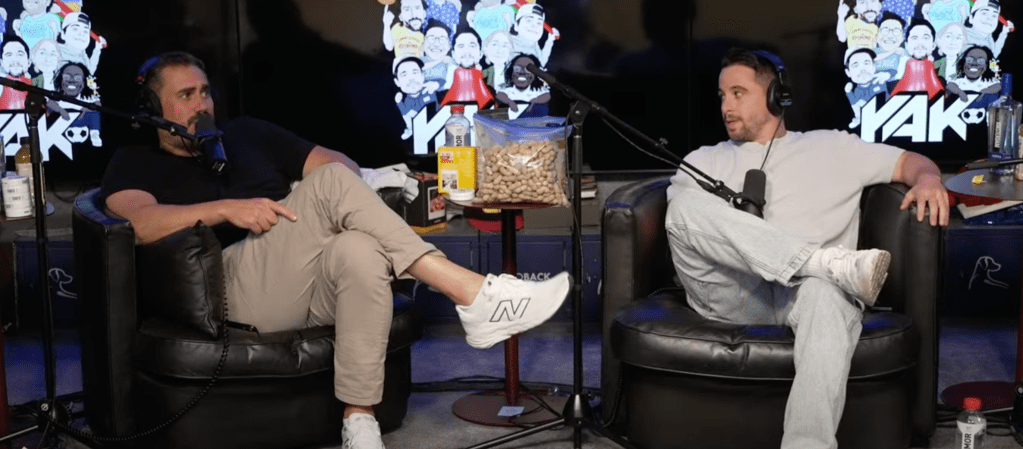 Two men in black armchairs talking into microphones with a bag of peanuts on a stand between them.